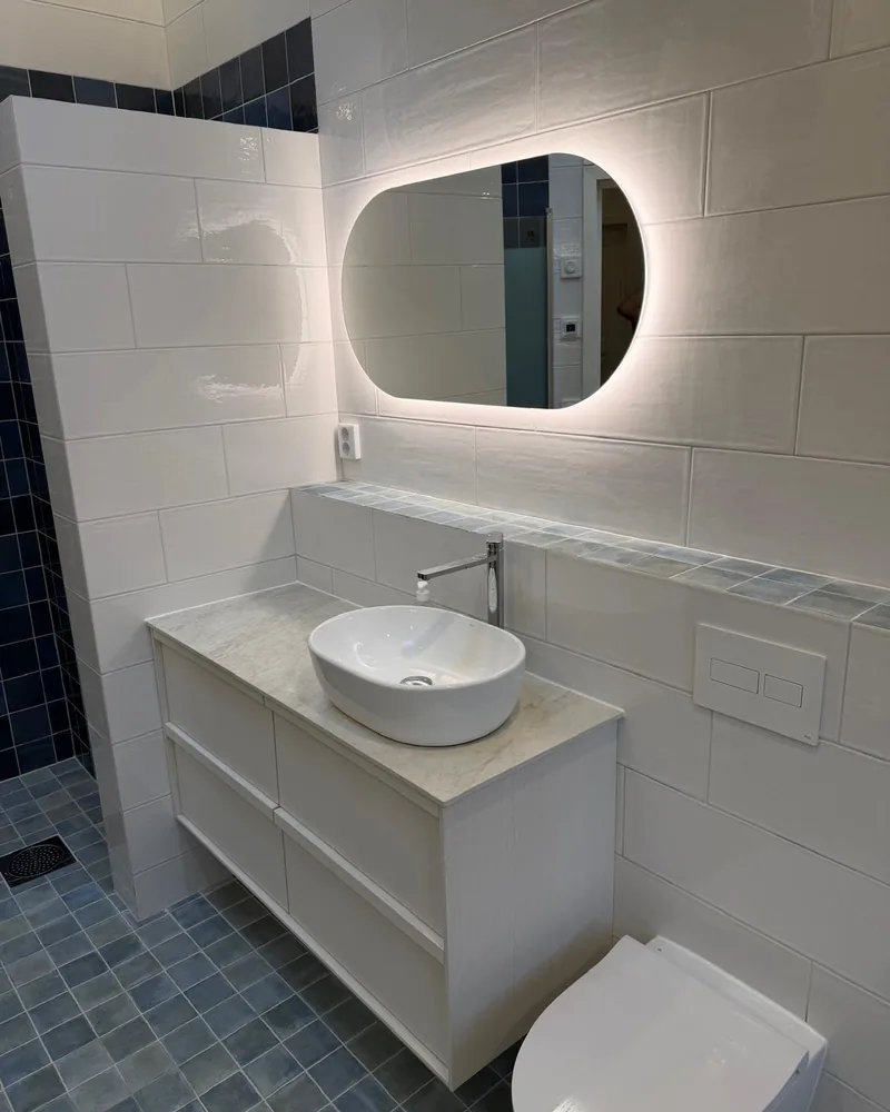Badrum renoverat av BHB Bygg AB med vitt kakel, blå mosaik och belyst oval spegel.